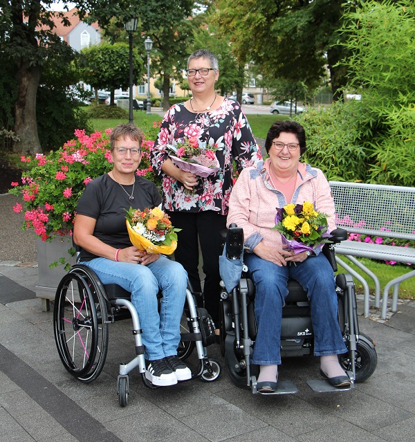3 Frauen, davon 2 im Rollstuhl, in einem sommerlichen Park. Jede Frau hält einen Blumenstrauß in der Hand.