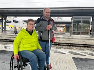 Eine Frau im Rollstuhl und ein Mann mit Langstock auf dem Bahnsteig.  Auf dem Boden ist ein Leitsystem zu erkennen.